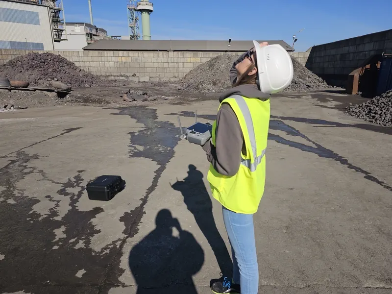 Drone pilot operating an industrial UAV over a coal and slag yard at a steel plant — bulk-pile aerial photogrammetry mission