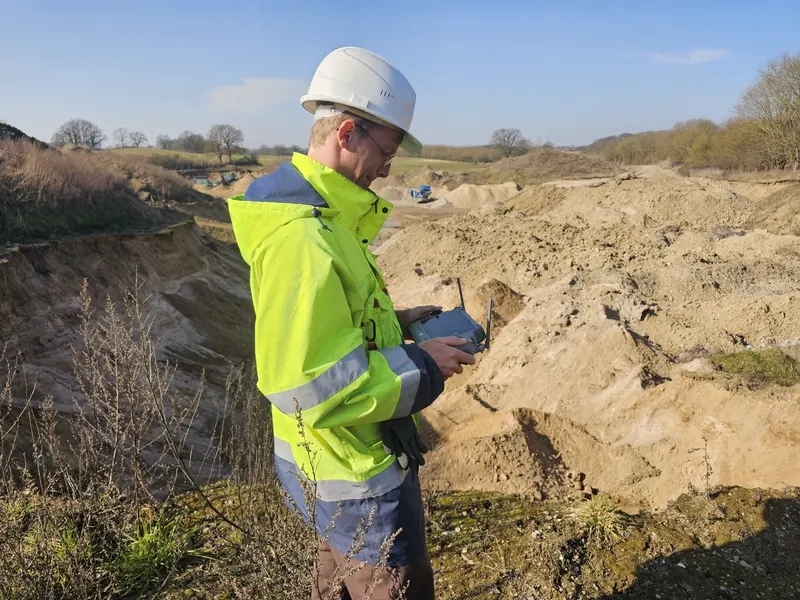 Sachtleben drone pilot in hi-vis vest and white hardhat operating a DJI controller above a sand quarry — RTK-corrected aerial survey