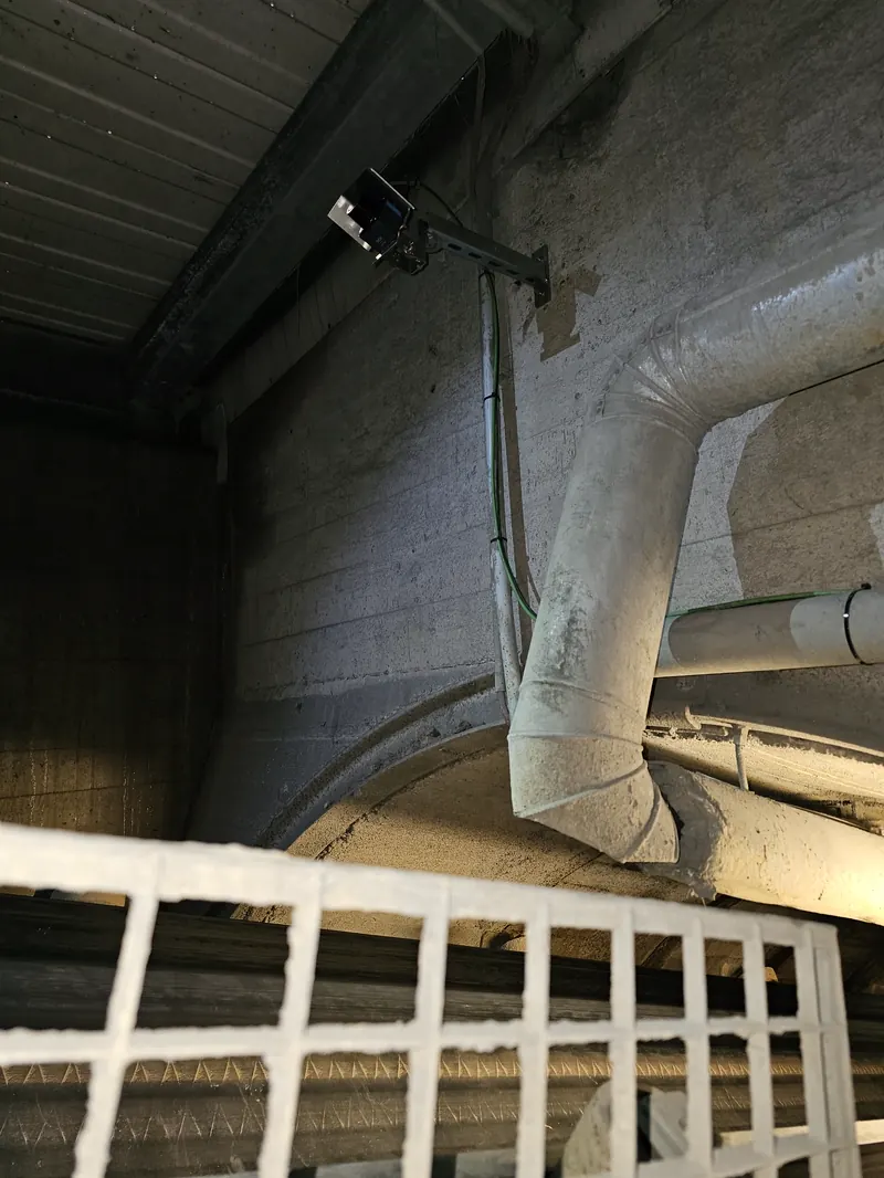 Cross-scan LiDAR sensor mounted under industrial bunker discharge — measuring volume flow leaving the silo onto the conveyor