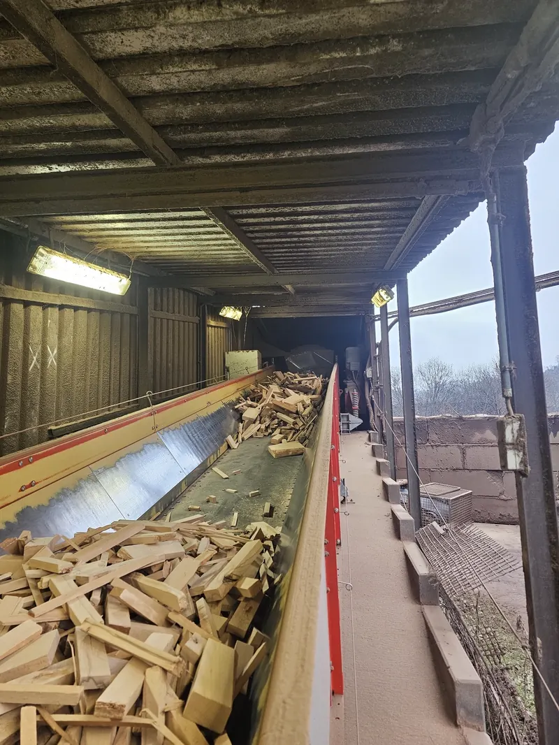 Wood chips streaming over an industrial conveyor belt under an OWL EYE® VOLUME FLOW 2D-LiDAR cross-scan unit — biomass and pellet plant